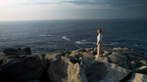 young caucasian woman standing on the rock while looking in the sea