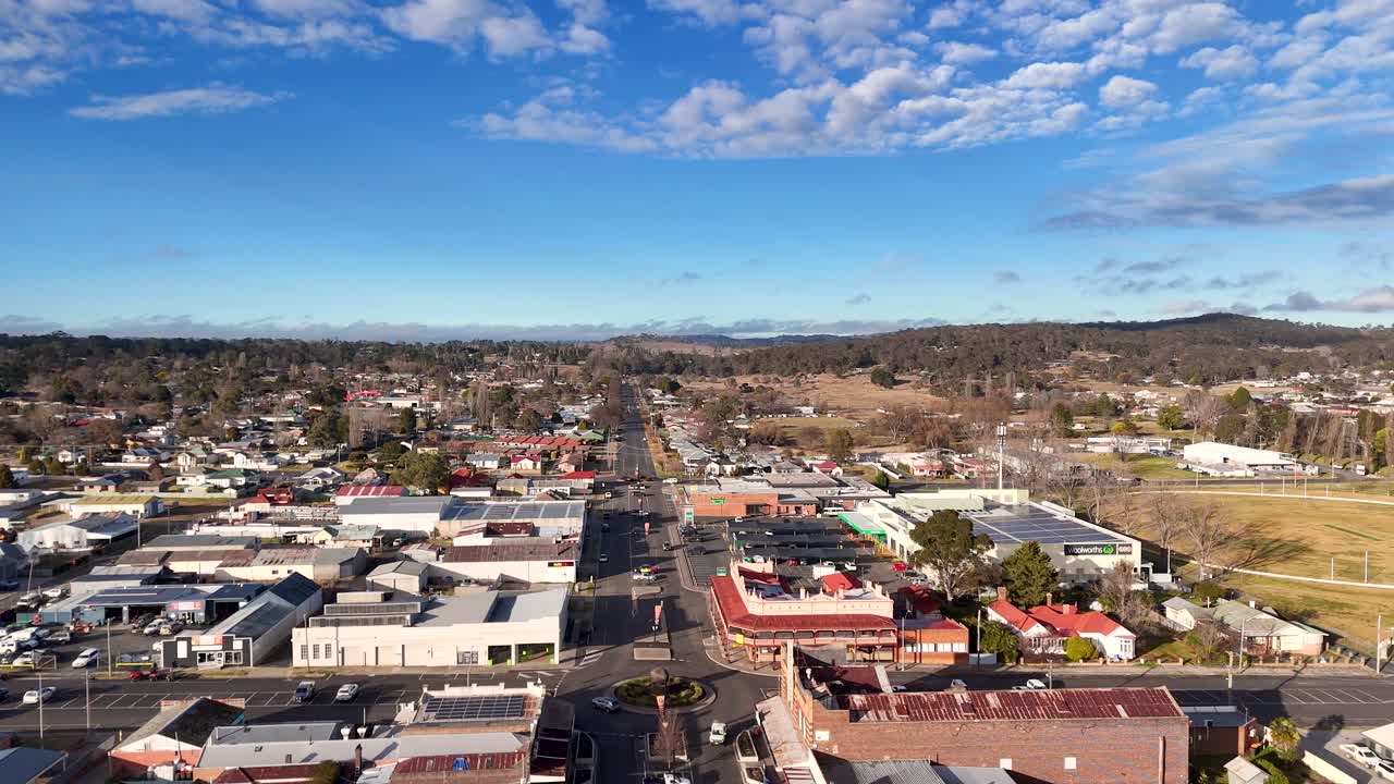 Drone camera glides above Glen Innes town center, revealing urban streets, commercial buildings, and residential areas under bright daylight with scattered clouds