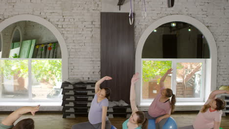 group of four active pregnant women and their female fitness trainer sitting on stability balls in gym and doing side stretching and torso rotations exercises