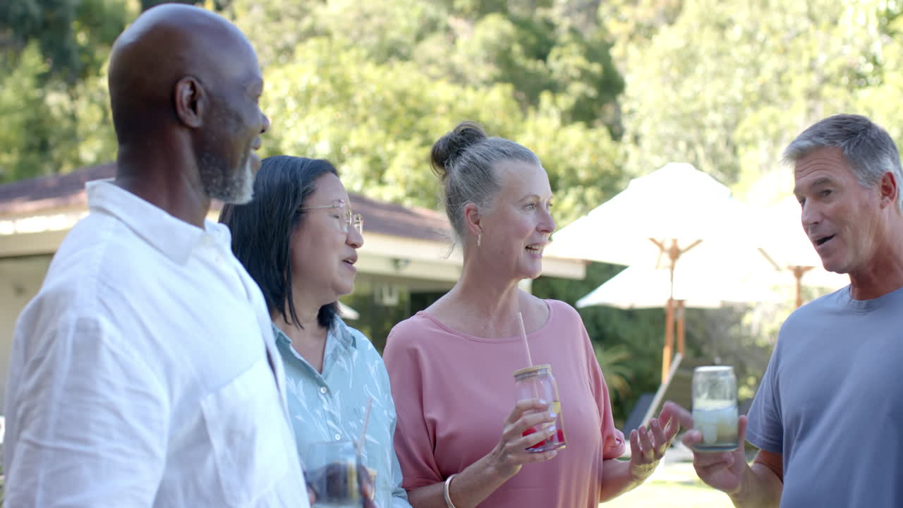 Senior diverse friends enjoying drinks and laughing together in garden in sunny garden