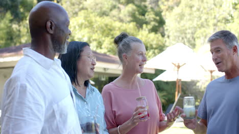 senior diverse friends enjoying drinks and laughing together in garden in sunny garden