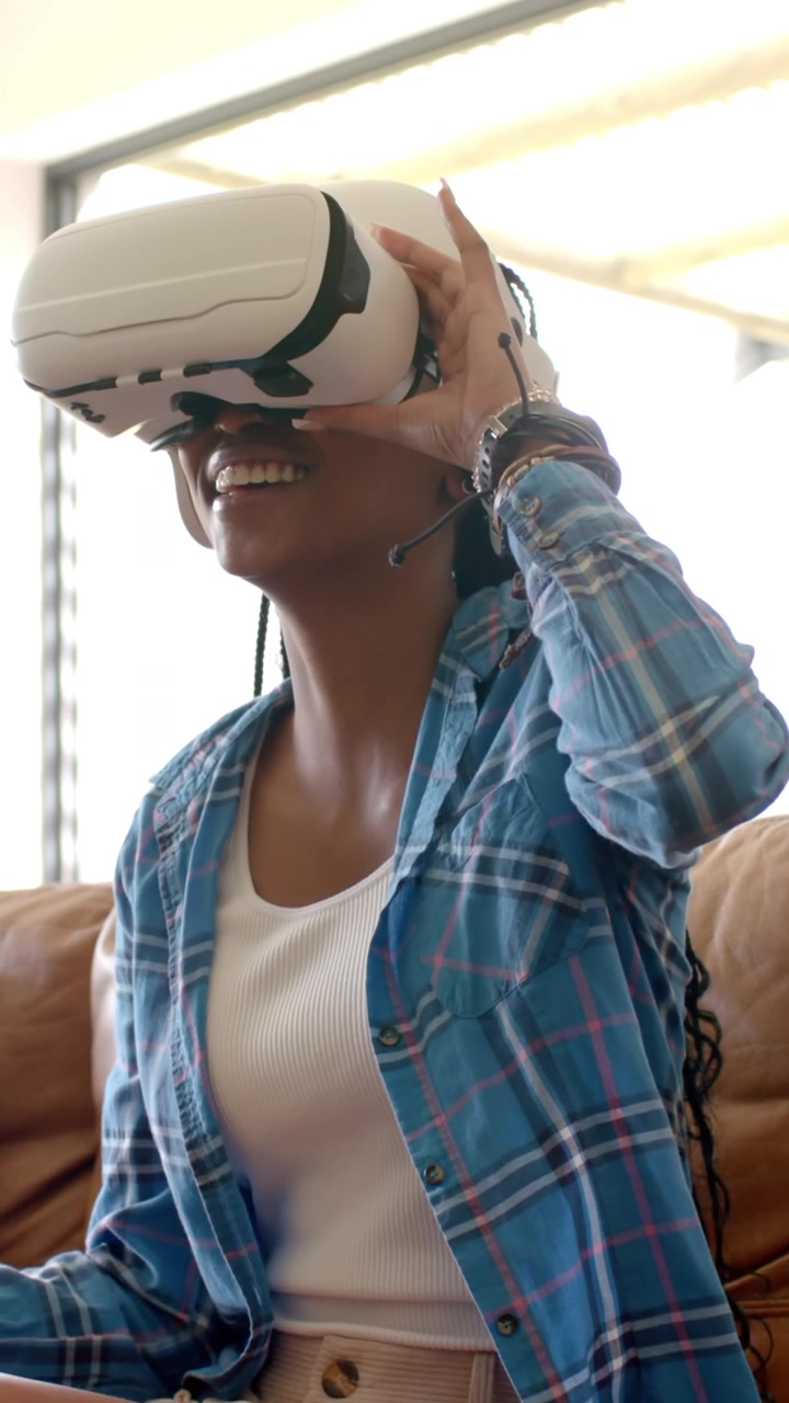 Video verticale: donna afroamericana sul divano vicino alla finestra che reagisce alla VR, regolando il visore per adattarlo
