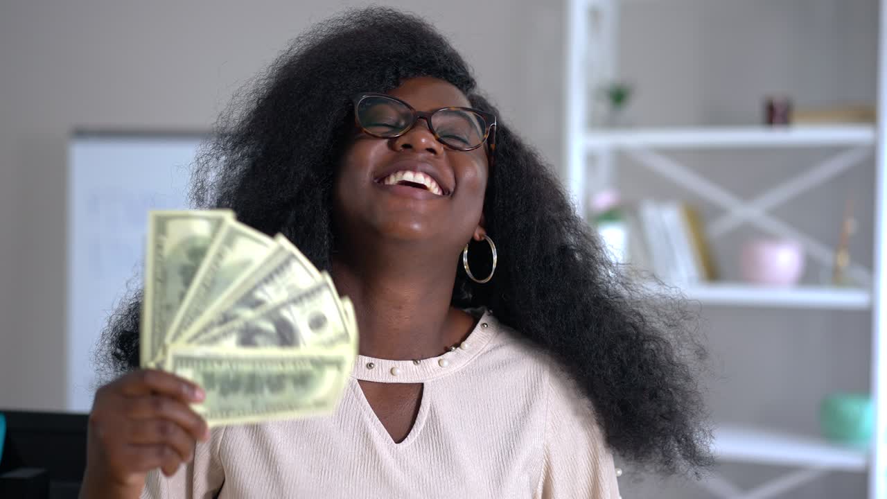 Portrait of happy wealthy African American young woman smiling boasting cash money indoors. Rich successful confident freelancer with income in home office. Slow motion.
