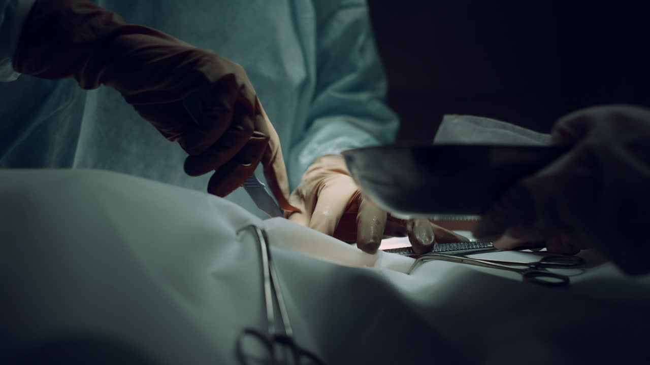 외과 의사의 손이 어두운 병원 병실에서 스칼펠을 사용하여 절개를 하고 있습니다.