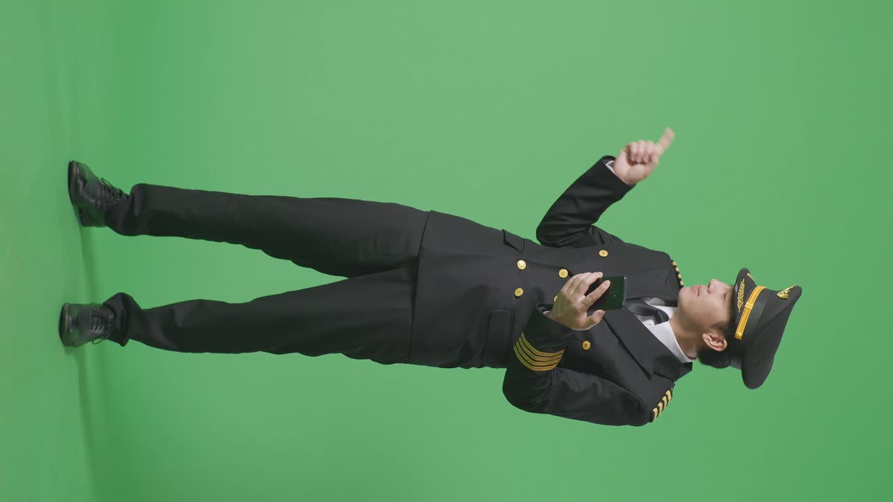 녹색 스크린 배경 스튜디오에 서 있는 동안 스마트폰을 사용하고 을 가리키는 아시아 남자 조종사의 전체 몸