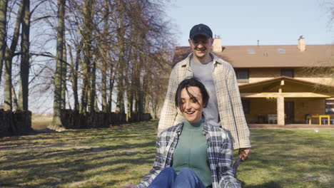 front view of caucasian man carrying his partner on a wheelbarrow in the countryside. their dog walks around them