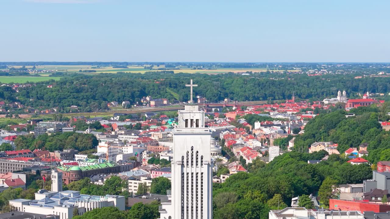 High-angle aerial view of the Church of the Resurrection in Kaunas, Lithuania, with its iconic tower rising above the colorful city and lush green surroundings on a clear summer day