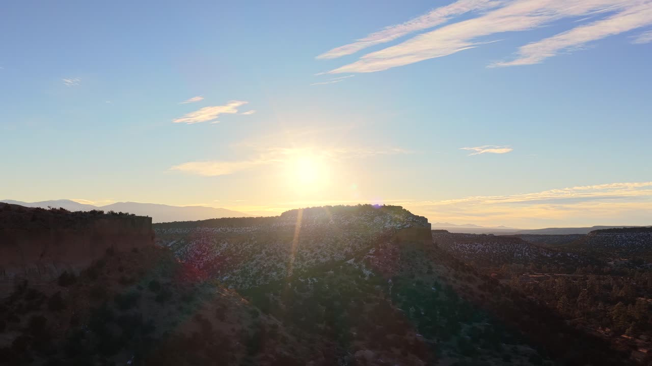 A dramatic drone shot captures a mesa silhouetted against the brilliant setting sun. High-contrast light defines the rugged cliffs and deep shadows of this high desert plateau landscape