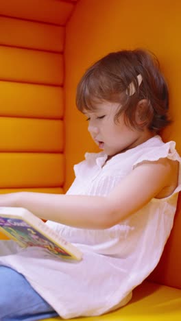 little girl seriously reads a book while sitting in a soft yellow chair, enjoying her free time at library - slow motion, vertical