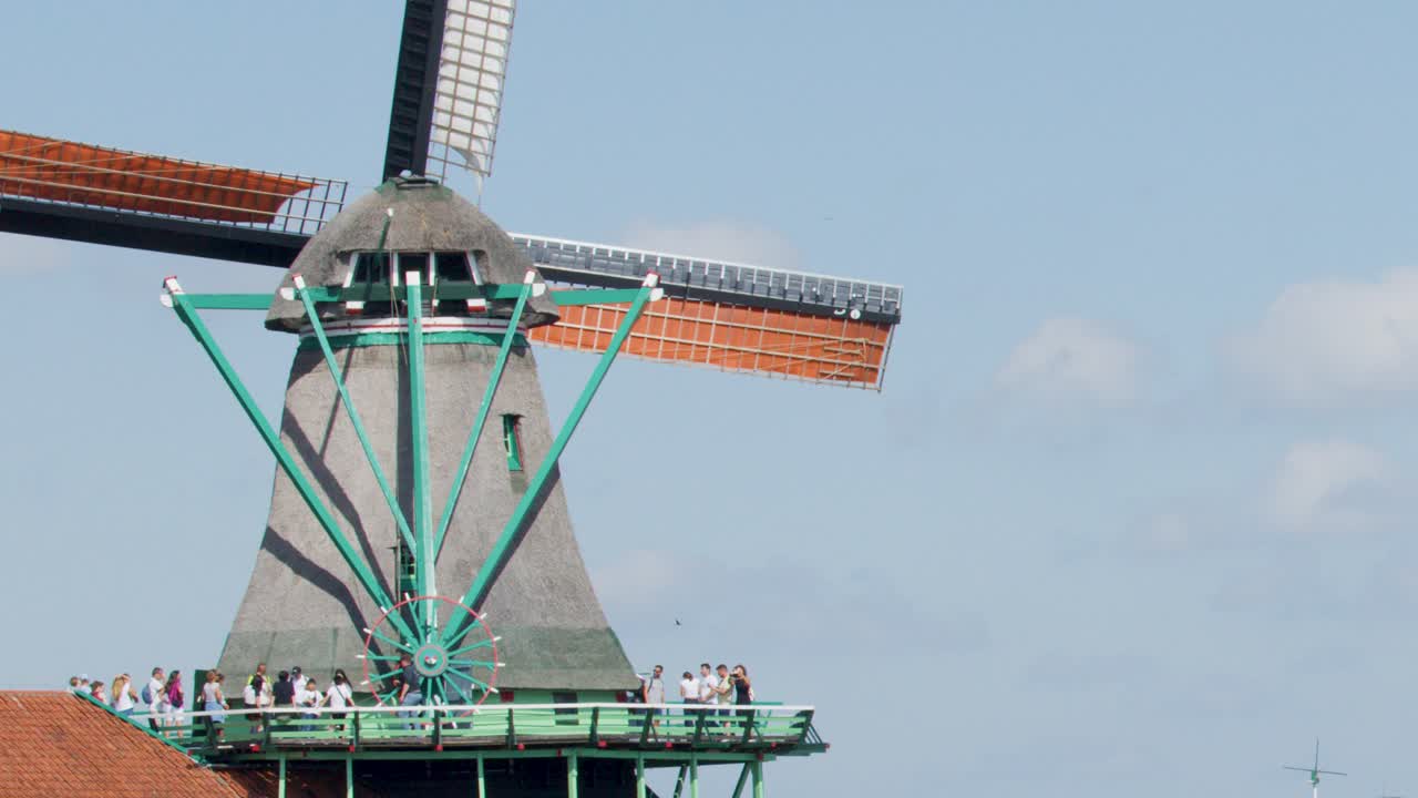 A classic Dutch windmill with green accents rotates slowly on a sunny day, observed by visitors on a viewing platform in Zaandam, Netherlands