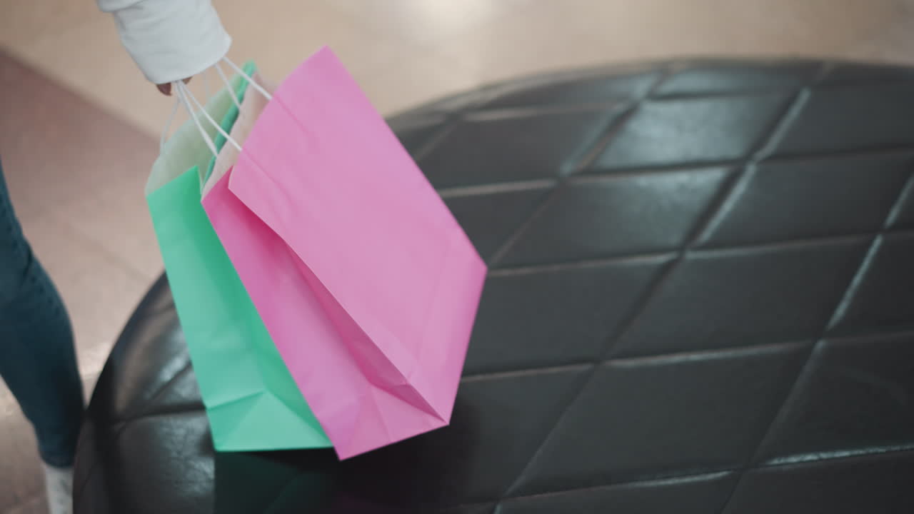 primer plano de una mano alcanzando para agarrar bolsas de compras coloridas de un asiento de cuero en un centro comercial bien iluminado, mostrando un ambiente de compras vibrante