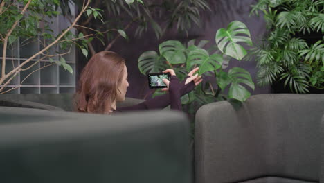 pretty woman sitting between cushioned chairs holding leaf with one hand and capturing photo on phone held in landscape mode, surrounded by lush indoor plants and soft ambient lighting