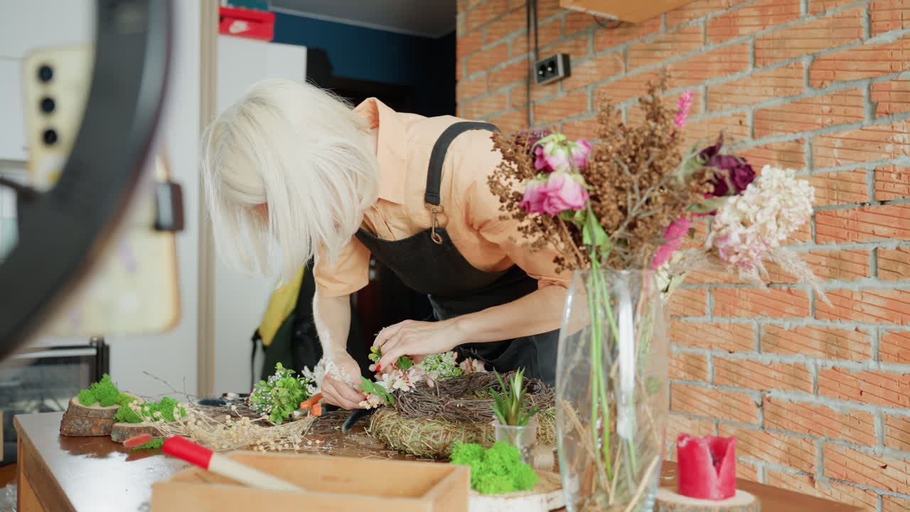 Female florist with blonde hair wearing apron working on floral wreath arrangement at home studio table surrounded by flowers, tools, and decor while recording creative process with lighting setup