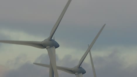 Dunkle-Wolken,-Die-Mit-Sanften-Sonnenuntergangsfarben-Hinter-Langsam-Rotierenden-Rotorblättern-Von-Windkraftanlagen-Im-Vordergrund-Vorbeiziehen