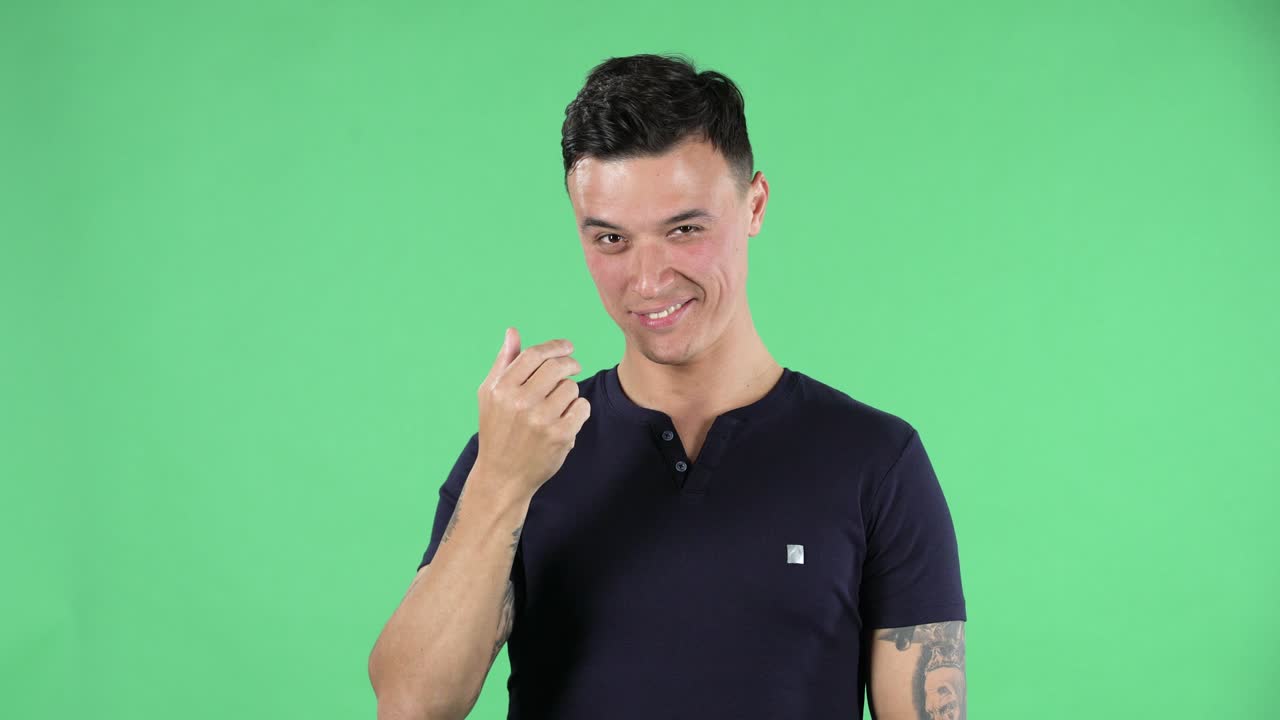 Portrait young guy is playfully waves his hand and shows a gesture to come here and winks. Burning brunette with stylish hair in a black T shirt and tattoos on his hands on a green screen in a studio