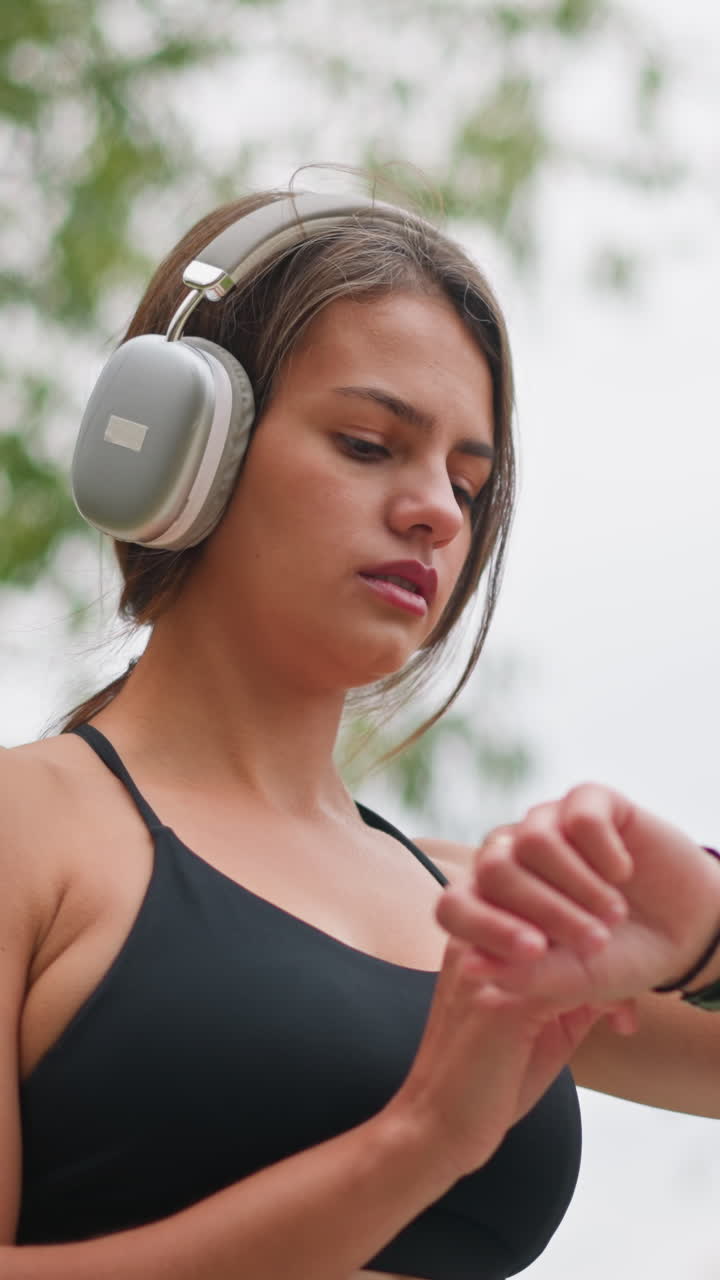 Portrait of focused young woman wearing black singlet and headphones, looking at her wristwatch as she checks time during outdoor fitness activity in a natural setting