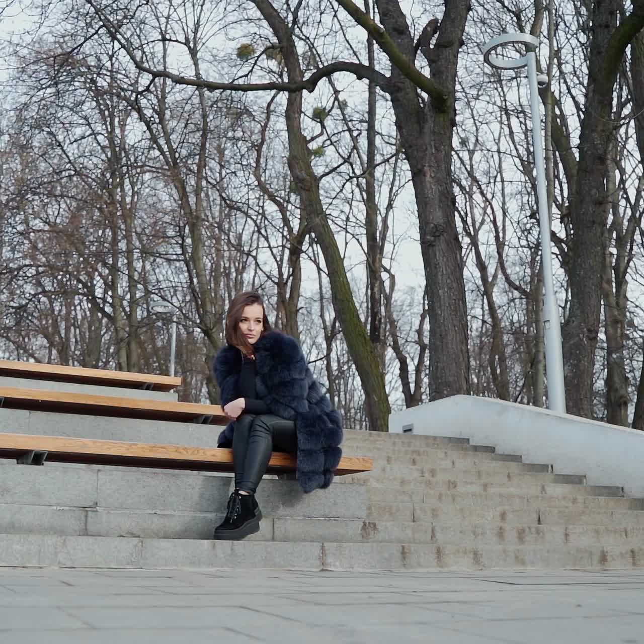 Girl sits on bench in the park. Beautiful young woman in fluffy coat sitting alone outdoors on the leafless trees background. View from below.