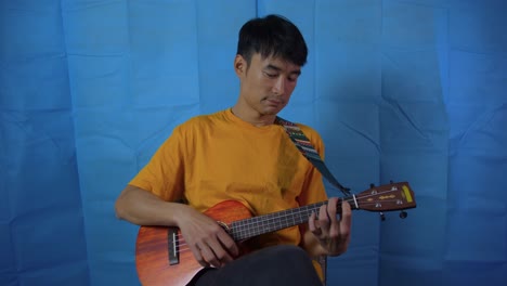 man slowly playing stringed ukulele musical instrument with blue background while seated