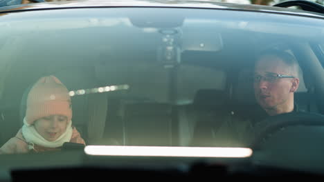 inside a car, as a man in a green jacket sits in the driver s seat, while his daughter, dressed in a pink beanie and jacket, smiles at him while focused on a smartphone in her hands