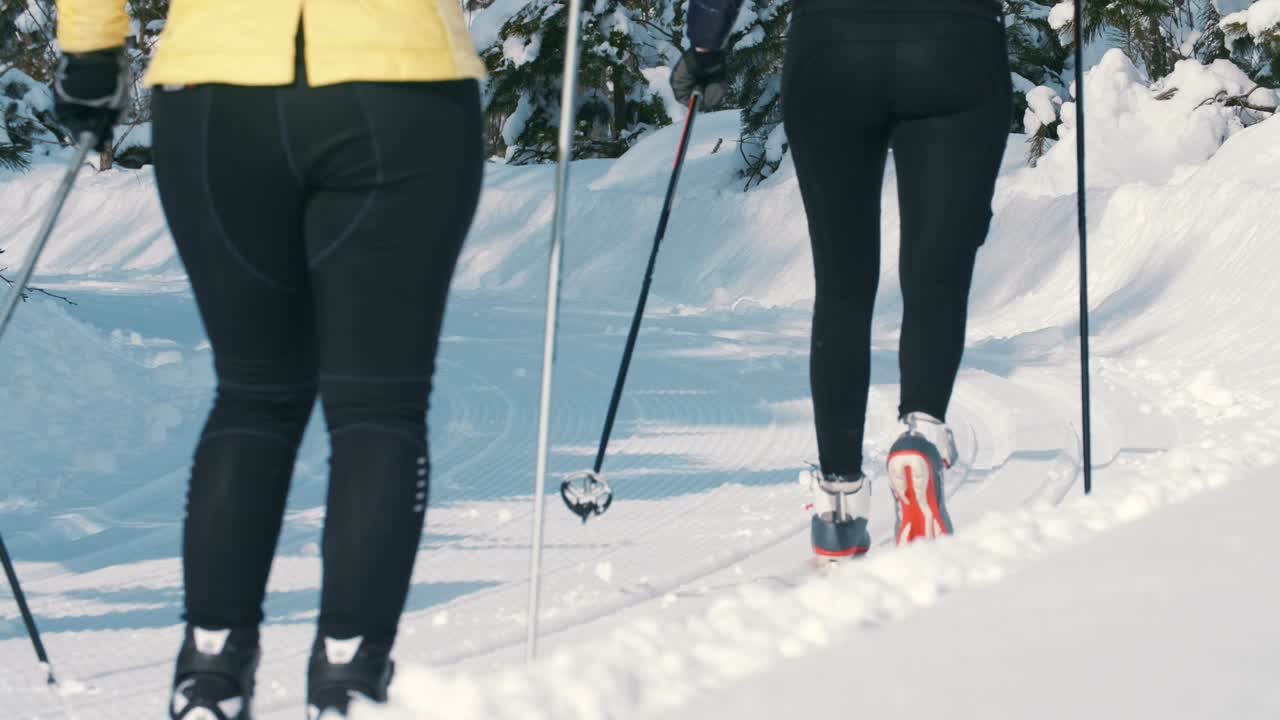 A ground-level, close-up shot of two cross-country skiers from behind, showcasing their technique and equipment as they glide along a groomed, sunlit snow trail in Kenauk, Quebec