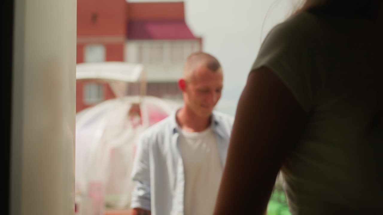 Hvidt par på tagterrasse-date: legesyg kvinde smiler nær gennemsigtig boble-pod, mand står bagved, urbant baggrundsmiljø med murstensbygning, varmt sollys, afslappet sommeroutfit, ægte terrasse-romantik
