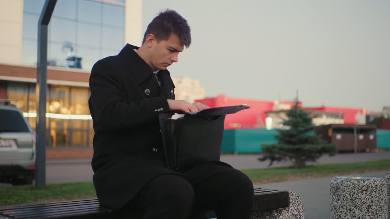 도시 환경에서 비즈니스 순간을 보여주는 도시 환경에서 현대적인 건물을 특징으로하는 배경과 함께 무언가를 찾는 브리프케이스를 통해 야외에 앉아있는 남자