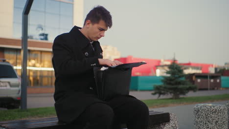 hombre sentado al aire libre revisando un maletín en busca de algo, con el fondo mostrando un edificio moderno en un entorno urbano, mostrando un momento de negocios en un entorno de ciudad