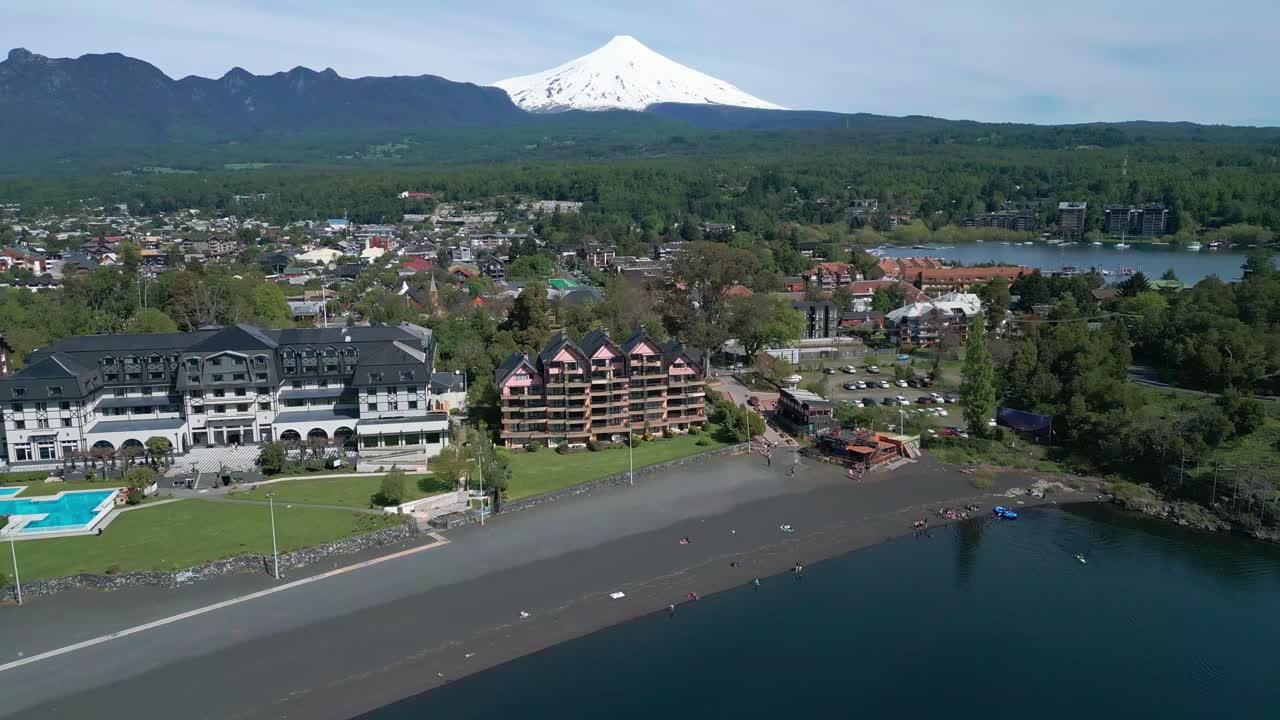Drone flying backward over beach, modern hotels, and black sand, revealing Pucón town and the Villarrica Volcano in the background