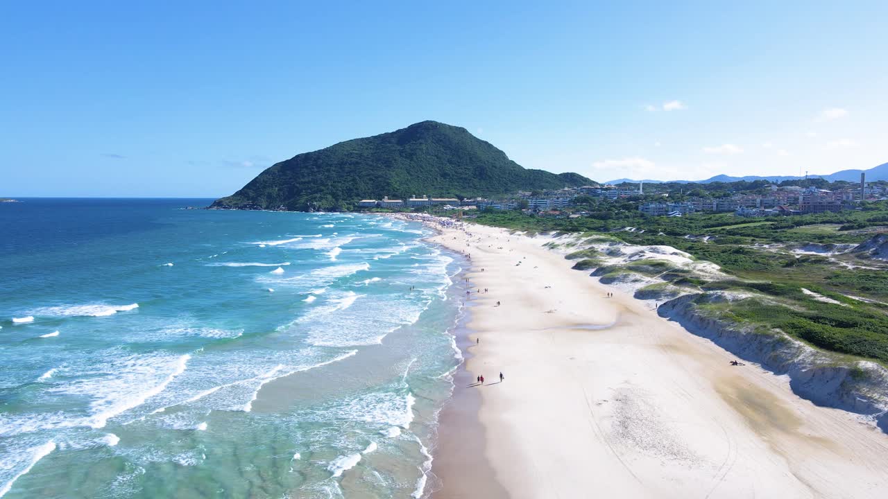 Ampia ripresa aerea cinematografica di Praia do Santinho a Florianopolis, Brasile, con onde turchesi dell'oceano, costa di sabbia bianca e il lussureggiante Morro das Aranhas sotto un cielo azzurro e limpido