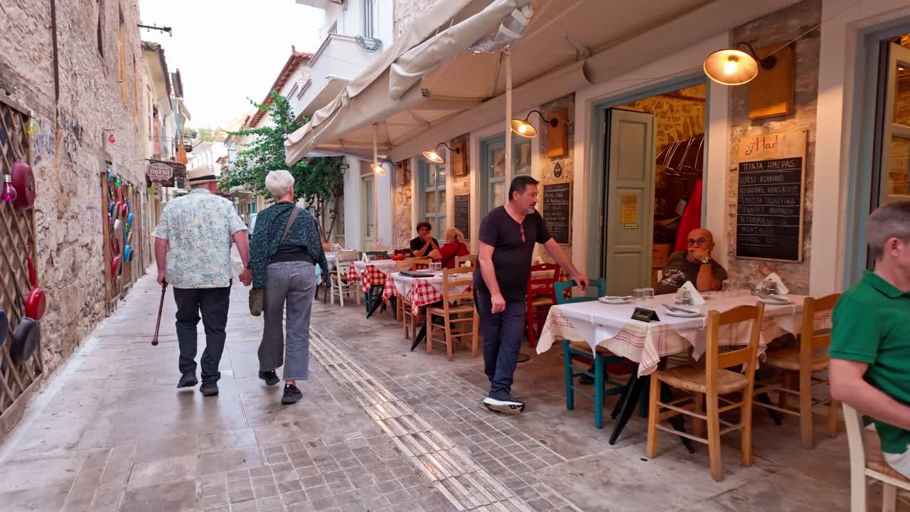 Diners enjoy Greek food al fresco city quaint streets, handheld walk shot