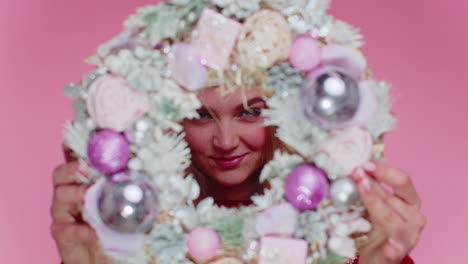 una mujer feliz lleva un suéter y un sombrero de navidad rojos, sostiene una corona festiva y se esconde detrás.