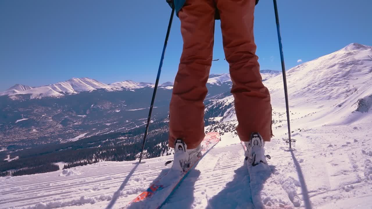 A slow motion low angle rear view of a skier going over a steep mountain cliff edge with the rocky mountains in the distance