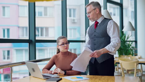 strict elderly businessman talks to a secretary. young woman sitting at her workplace near the window and listens to senior entrepreneur.