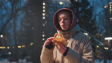 young woman standing in park on winter evening, eating snack wrapped in paper while enjoying nature, urban setting with snow-covered ground and street lights in background