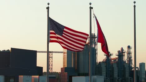 a usa and arkansas flag wave in the wind at sunset with a rice manufacturing plant in the background