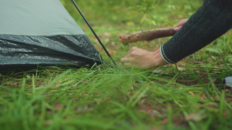 close up leg view showing companion standing beside crouched camper while he hammers tent peg into soft forest ground amid grass securing camp shelter for outdoor adventure