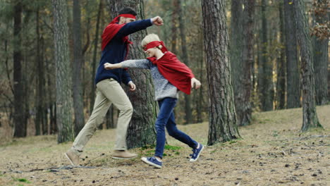 padre e hijo caucásicos alegres con capas rojas corriendo en el bosque y jugando a ser superhéroes