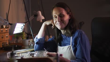 caucasian female jeweller sitting at desk, making jewelry and smiling at camera