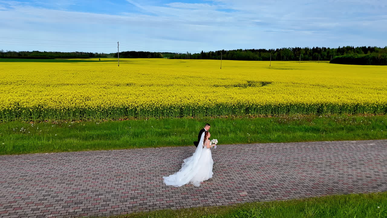 Et nygift par slentrer langs en brostensbelagt gade ved siden af en gul mark, mens en jævn drone følger dem og fremkalder romantik, engagement og pastoral ro efter ceremonien