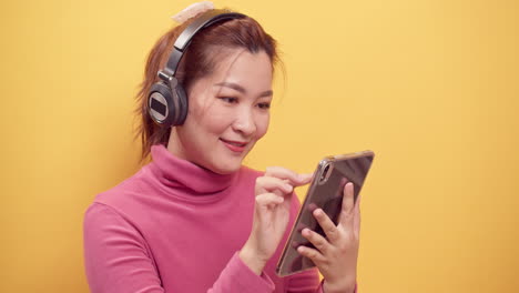 smiling asian woman using a digital tablet and streaming application online listening to music relaxations with headphones on bright yellow background