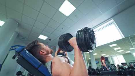 Side view of a strong man with biceps lying on desk and lifting dumbbells in gym. Muscular man doing workout on the background of sport center. Pretty woman looks at bodybuilders training.