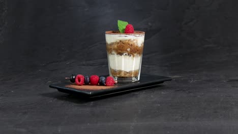 a glass of tiramisu garnished with berries, mint and cocoa powder is presented on a spinning plate