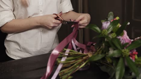 A-girl-in-a-white-shirt-cuts-long-pink-ribbons-and-purple-broad-ribbons-of-guipure-to-decorate-a-bouquet-of-flowers-on-a-table.-Close-up