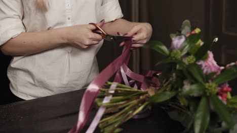 a girl in a white shirt cuts long pink ribbons and purple broad ribbons of guipure to decorate a bouquet of flowers on a table. close up