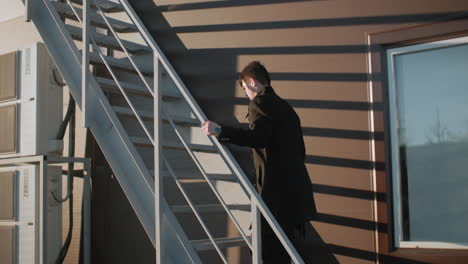 investor in black coat climbing staircase, holding iron railing and briefcase in right hand, with sunlight casting shadow on modern striped building wall