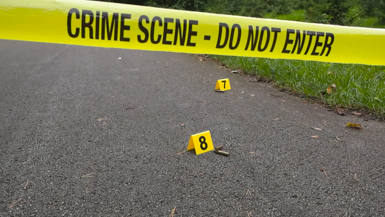 Yellow colored police line crime scene tape in front of numbered Crime markers that are placed next to high caliber rifle or weapon bullets or ammunition on a ashpalt road. Bokeh grassy background