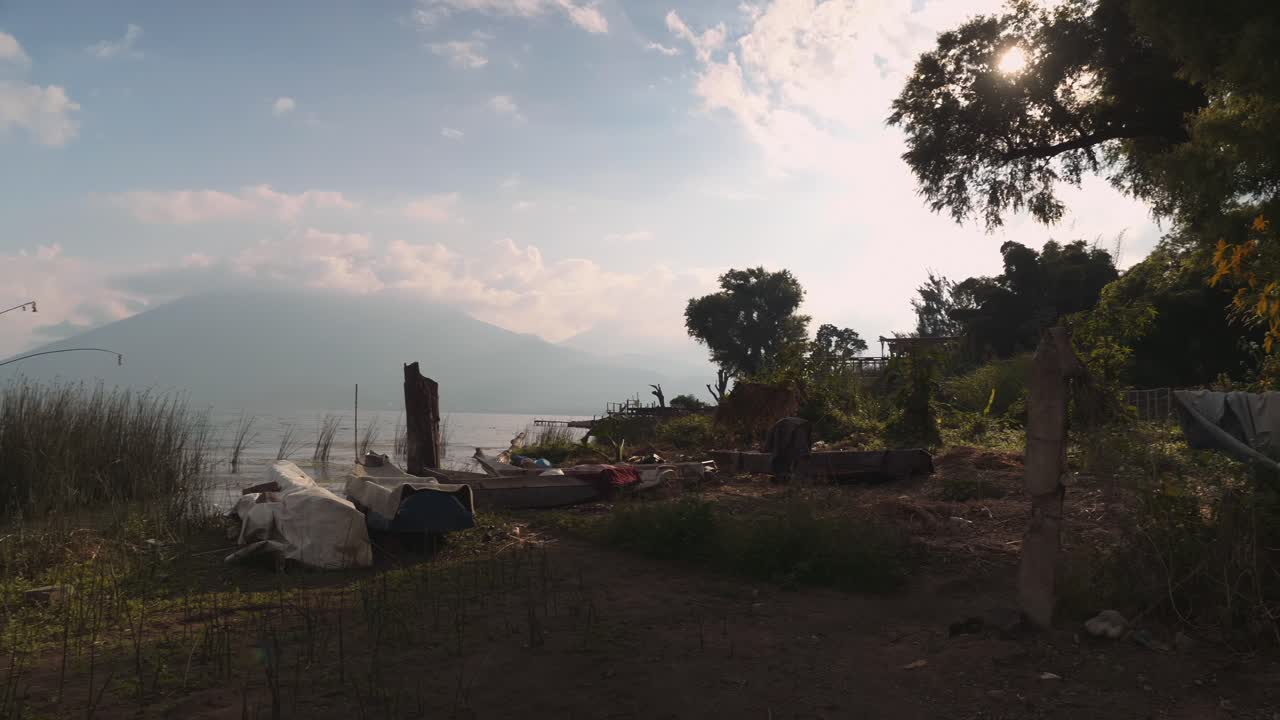 Små både ved bredden af Atitlan-søen ved solopgang med en skybeklædt vulkan i baggrunden