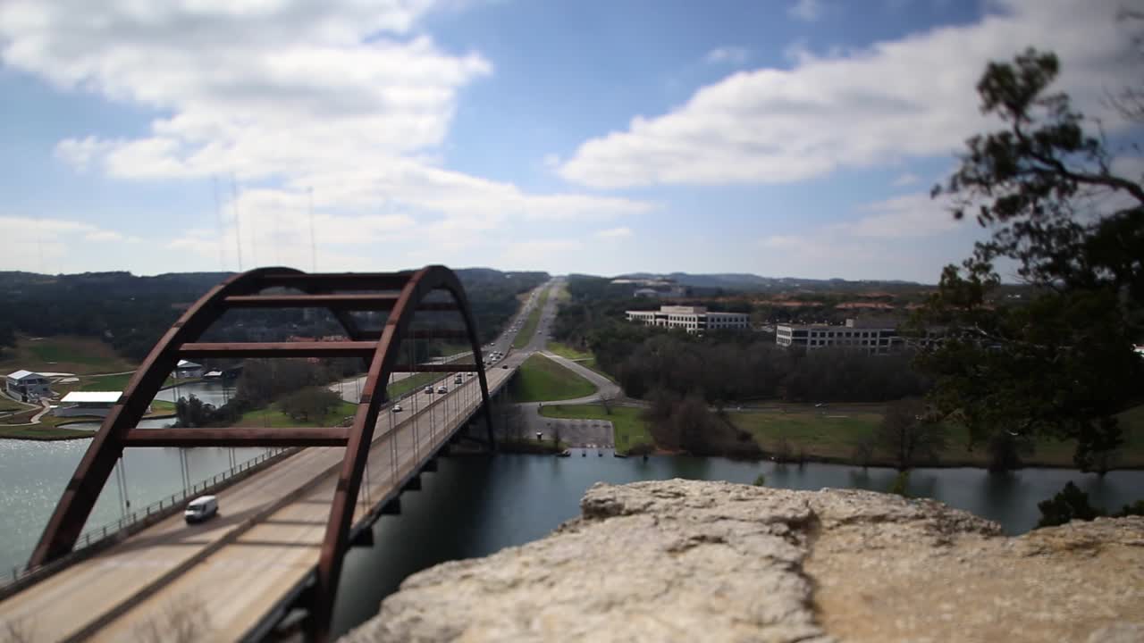 Austin Pennybacker Bridge tilt-shifted time-lapse, focus isolated on center of bridge and boat ramp across the river, sun hits the bridge and then continues to green field across the river.