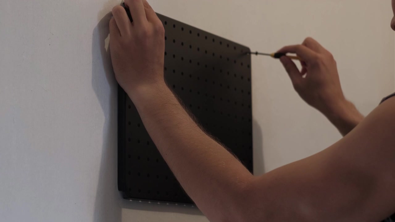 Medium shot of hands installing black pegboard on wall using screwdriver indoors for home improvement and workspace organization.
