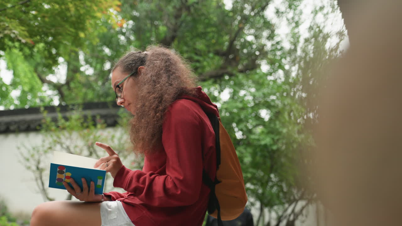 Signora con felpa con cappuccio assorta in un libro vicino alla vegetazione. Giovane donna con felpa rossa con cappuccio che sfoglia pagine su una panchina del parco. Scatto spontaneo di donna con capelli ricci e zaino immersa nella lettura all'aperto.